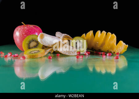 Leckere und gesunde Snack - frische Banane, in Scheiben geschnittene grüne Kiwi, Apfel, Hawaiianische Ananas am grünen Tisch - Schwarzer Hintergrund Stockfoto