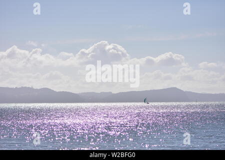 Lila und violetten Reflexen am Meer im Gegenlicht mit Segel Yacht und graue Silhouette der Hügel im entfernten Hintergrund. Stockfoto