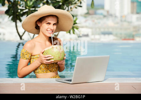 Porträt der glückliche junge Frau in Stroh Hut stehen am Pool mit geöffneten Laptop auf und trinken Coconut cocktail Stockfoto