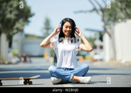 Portrait von fröhlichen Vietnamesischen Jugendmädchen sitzen auf der Straße mit ihr Skateboard und das Hören von Musik in großen Kopfhörer Stockfoto