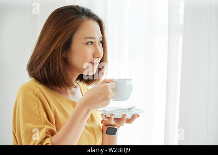 Schönen lächelnden jungen vietnamesischen Frau trinkt Tasse Cappuccino und schauen durch das Fenster Stockfoto