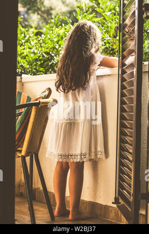 Baby Mädchen in weißem Kleid und mit langen braunen Haaren, stehend auf einem Balkon auf den Garten Hintergrund. Stockfoto