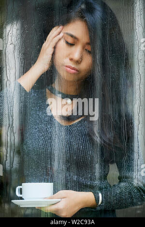 Traurig Asiatische junge Frau, die in der Nähe des Fensters und halten Tasse Kaffee regnet hinter dem Fenster und schaut sie umgekippt Stockfoto