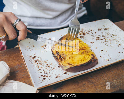 Hände mit Messer und Gabel Käse Toast aus weisser Keramik Platte auf hölzernen Tisch im Café. Stockfoto