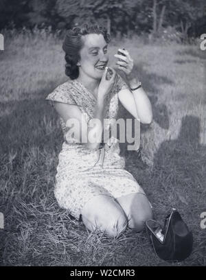 1950, historische, eine junge Dame in einem Kleid nach unten außen Kniend auf einem etwas Gras mit einem kleinen Pocket Mirror ihren Lippenstift, England, UK. Stockfoto