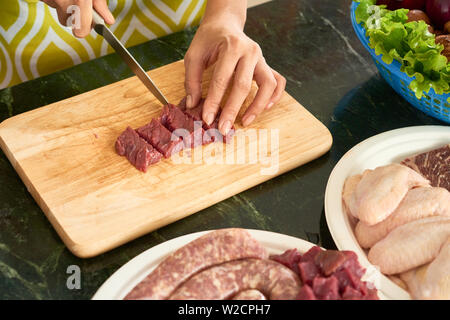 In der Nähe von Frau Schneiden von rohem Fleisch in kleine Stücke mit Küchenmesser auf schneidebrett am Tisch Stockfoto