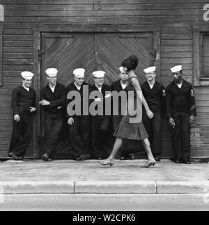 Mode in den 60er Jahren. Eine Gruppe von amerikanischen Matrosen stehen bei den Docks auf Landgang und macht eine schöne Ergänzung zu den Fashion Shooting. Ein junges Modell leitet Sie trägt ein Kleid mit passender Tasche und Schuhe. Schweden 1964 ref BV 59-5 Stockfoto