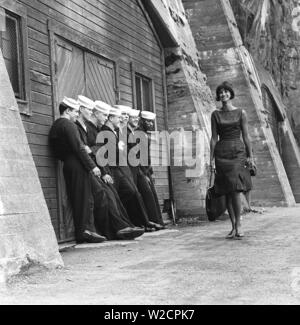 Mode in den 60er Jahren. Eine Gruppe von amerikanischen Matrosen stehen bei den Docks auf Landgang und macht eine schöne Ergänzung zu den Fashion Shooting. Ein junges Modell leitet Sie trägt ein Kleid mit passender Tasche und Schuhe. Schweden 1964 ref BV 59-6 Stockfoto