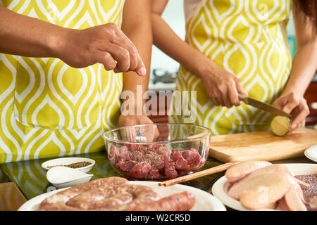 Close-up des Menschen hinzufügen Pfeffer in Stücke von Fleisch in der Schüssel, während Frau schneiden Gemüse an Bord, stehend in der Schürze in der Küche Stockfoto