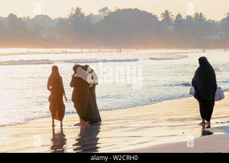 Gruppe von jungen muslimischen Frauen am Strand bei Sonnenuntergang, eine reife Frau mit Taschen kommt vorbei und schaut sie an. Weligama, Sri Lanka. Stockfoto