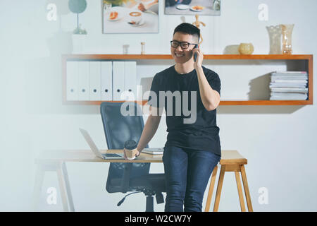 Asiatische junge Unternehmer in Freizeitkleidung in der Nähe von seinem Arbeitsplatz Gespräch am Handy und Lachen im Büro Stockfoto