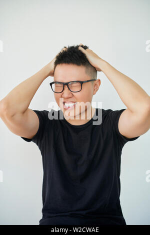 Portrait von Asiatischen jungen Mann in Brillen seinen Kopf mit den Händen festhalten mit hetzerischen Gesichtsausdruck auf weißem Hintergrund Stockfoto