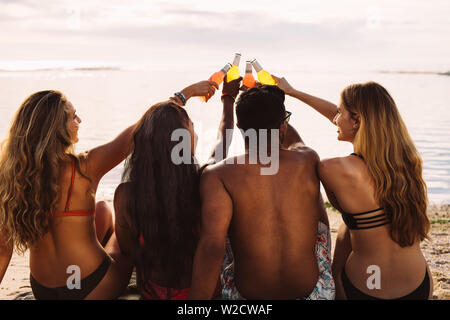 Ansicht von hinten von einer Gruppe von Freunden zusammen sitzen am Strand toasten Flaschen alkoholfreien Getränk. Mann und Frau eine tolle Zeit zusammen in einer Ferienwohnung in der Nähe von Stockfoto