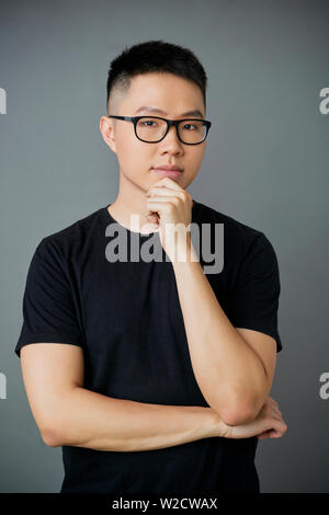 Portrait von Asiatischen jungen Mann in Brillen und in schwarzen T-Shirt auf Kamera schaut mit Zuversicht Ausdruck auf grauem Hintergrund Stockfoto