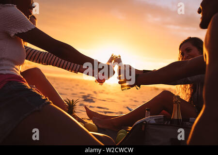 Freunde eine Party in der Nähe der Strand bei Sonnenuntergang sitzen. Lächelnd Freunde auf einen Urlaub toasten Getränke sitzen am Strand mit Sonne in den Hintergrund. Stockfoto