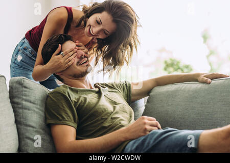 Junge Frau hinter ihren Freund auf dem Sofa stehen und die seine Augen zu Hause. Frau überraschend Mann zu Hause. Stockfoto