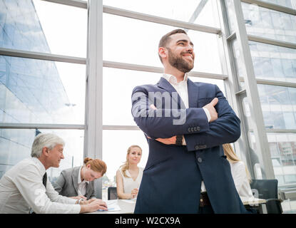Zuversichtlich, dass junge Unternehmer Stehen mit verschränkten Armen lächelnd, Team von seinen Kollegen an dem Tisch im Hintergrund sitzen Stockfoto