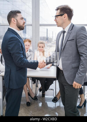 Handshake von Geschäftspartnern nach Diskussion der Finanzberichte, ihr Team im Hintergrund Stockfoto