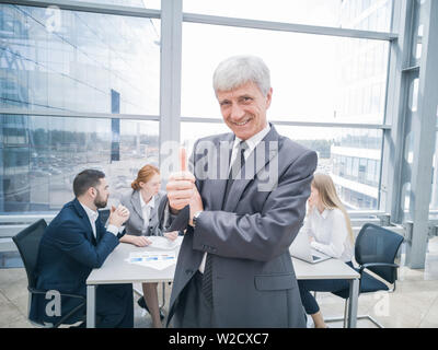 Zuversichtlich reife Geschäftsmann, Daumen hoch und Lächeln, Team von seinen Kollegen an dem Tisch im Hintergrund sitzen Stockfoto