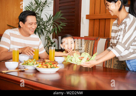 Asiatische glückliche Familie am Tisch sitzen und essen zusammen mit Mutter mit Salat und lächelnd Stockfoto