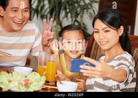 Asiatische junge Mutter holding Handy und Selfie portrait Ihrer Familie, während Sie Essen am Tisch Stockfoto