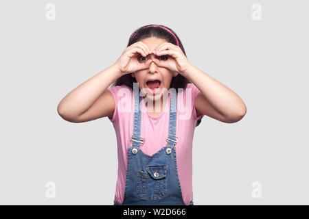 Portrait von überrascht junge Mädchen in rosa t-shirt und blauen Overalls stehen mit dem Fernglas Geste die Hand auf die Augen und suchen mit entsetzten Gesicht. indoor s Stockfoto