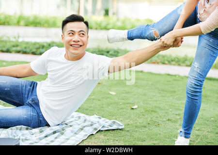 Asiatische lächelnden Mann, der versucht, sich von der Gras mit Hilfe der jungen Frau zu stehen nach dem Picknick im Park Stockfoto
