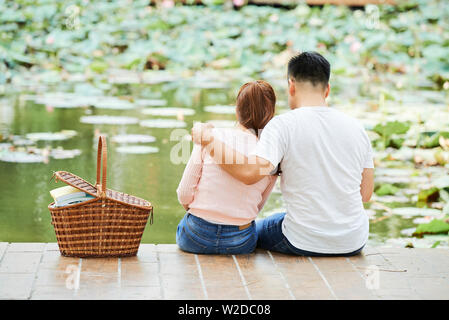 Ansicht der Rückseite des jungen Paares auf Pier in der Nähe des wunderschönen Sees und umarmen einander mit Picknick Korb in der Nähe von Ihnen Stockfoto