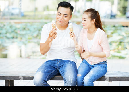 Asiatisches junges Paar in den weißen T-Shirts und Jeans sitzt auf der Bank und genießen Eis zusammen im Freien Stockfoto