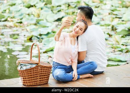 Gerne asiatische Frau, die Selfie portrait von sich selbst auf Mobiltelefon von Pier in der Nähe des Teiches zusammen mit ihrem Freund Stockfoto