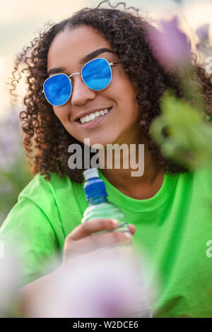 Gemischte Rasse schöne Afrikanische Amerikanische biracial Mädchen Teenager weibliche junge Frau trägt blaue Sonnenbrille und trinken eine Flasche Wasser im Bereich der Pin Stockfoto