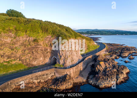 Black Arc Tunnel und Causeway Coastal Route. Malerische Straße entlang der östlichen Küste des County Antrim, Nordirland, Großbritannien. Luftaufnahme im Sonnenaufgang Licht Stockfoto