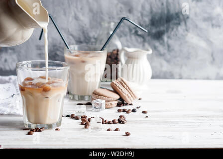 Gießen Milch von Krug Glas mit gefrorenen Kaffee Eiswürfel auf weißem Holz- Hintergrund und graue Betonwand, selektiver Fokus, Tageslicht Stockfoto