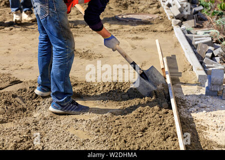 Der Arbeitnehmer richtet die Stiftung mit Sand und eine Schaufel unter der hölzernen Ebene für die Verlegung von Platten auf dem Hintergrund der Website für Arbeit vorbereitet Stockfoto