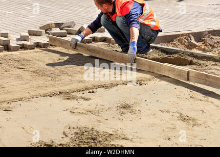Auf dem Bürgersteig, ein Arbeiter richtet die sand Plattform mit einem Holzbrett, bereitet die Grundlage für die Festlegung der Pflastersteine. Stockfoto
