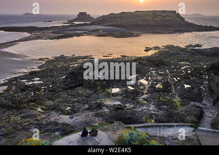 Zwei jüngere Männer sitzen auf einem Felsen watchig den Sonnenuntergang von St Malo, Bretagne, Frankreich Stockfoto