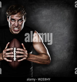 Fußballspieler Porträt holding American Football starrte aggressiv an der Kamera auf Schwarze Schiefertafel Hintergrund mit Kopie Platz für Text oder Design. Kaukasische männliches Modell in seiner 20. Stockfoto