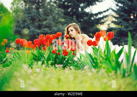 Schöne junge Frau mit hübschen lächelndes Gesicht in Blumenbeet mit roten Tulpen auf grünem Gras in Park liegen im Freien Stockfoto