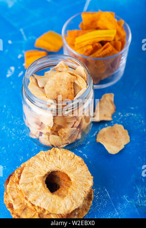 Apfel, Ananas und Mango Chips in einem Glas und Stücke dieser Früchte auf einem Tisch Stockfoto