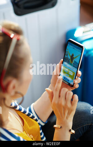 Über die Schulter geschossen von Jungen Solo touristische Frau in gestreifte Jacke in der Nähe der Couch und Trolley Taschen bei modernen Haus im sonnigen heißen Sommertag Sitzung buchen Fl Stockfoto