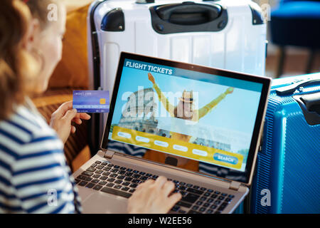 Über die Schulter geschossen von angesagten Solo touristische Frau in gestreifte Jacke mit Kreditkarte buchen Flüge online auf einem Laptop in der Nähe der Couch und tro Sitzen Stockfoto