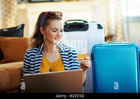 Elegante solo touristische Frau in gestreifte Jacke mit Kreditkarte kaufen Flüge online auf einem Laptop beim Sitzen in der Nähe der Couch und Trolley Taschen bei modernen Ho Stockfoto