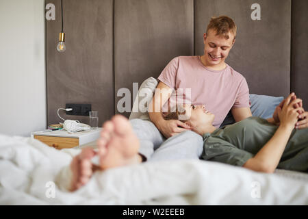 Glückliches junges Ehepaar unterhalten, während sie sich auf dem Bett am Wochenende morgens Stockfoto