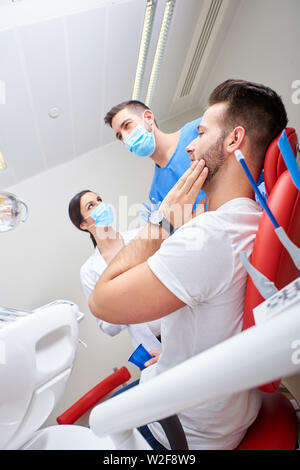 Porträt von einem gutaussehenden jungen Patienten in den Schmerz mit dem Zahnarzt und seinem Assistenten. Stockfoto