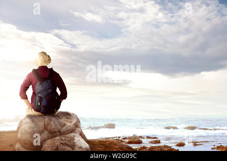 Ansicht der Rückseite des asiatischen Mann in Hut mit Rucksack sitzen auf den Felsen und mit Blick auf das Meer Blick auf den Strand Stockfoto