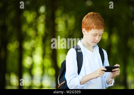 Redheaded Schüler spielen mit seinem Handy auf dem Weg nach Hause in den Park nach dem Unterricht in der Schule Stockfoto