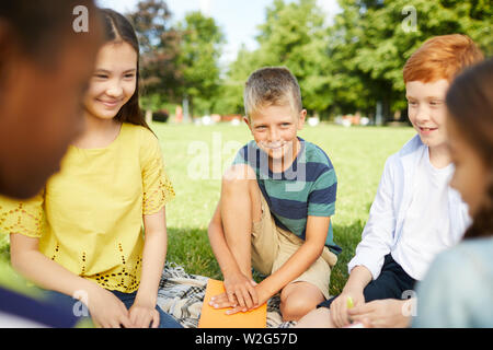 Fünf lächelnde Schüler im Kreis außerhalb auf Gras im Park sitzen und spielen lustige Spiele Stockfoto