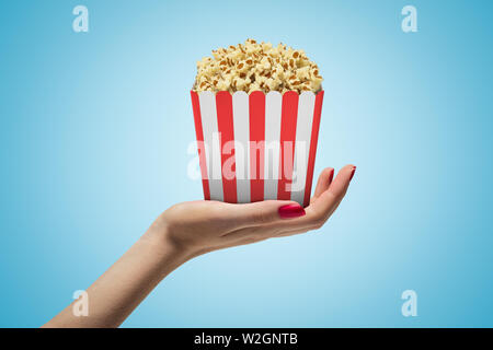 Seite Nahaufnahme des Woman's Hand nach oben gedrückt und gestreifte popcorn Box auf hellblauem Hintergrund Farbverlauf. Stockfoto