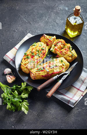 Gefüllte Zucchini mit Tomaten, Zwiebeln und Käse auf schwarzem Teller mit Messer und Gabel serviert, vertikale Ansicht von oben Stockfoto