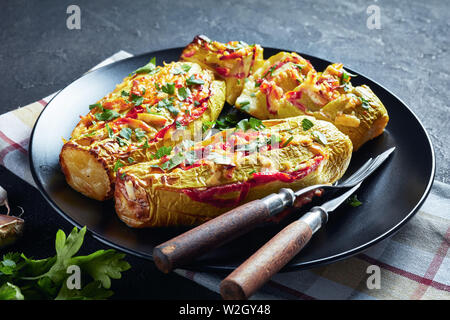 Gefüllte Zucchini mit Tomaten, Zwiebeln und Käse auf schwarzem Teller mit Messer und Gabel auf einen konkreten Tisch serviert, horizontale Ansicht von oben, Clo Stockfoto